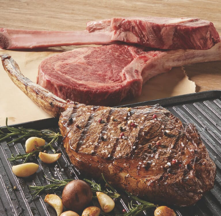 A seasoned, grilled Cowboy Ribeye and garlic and rosemary sprigs on a black platter, with two steaks waiting to be cooked.