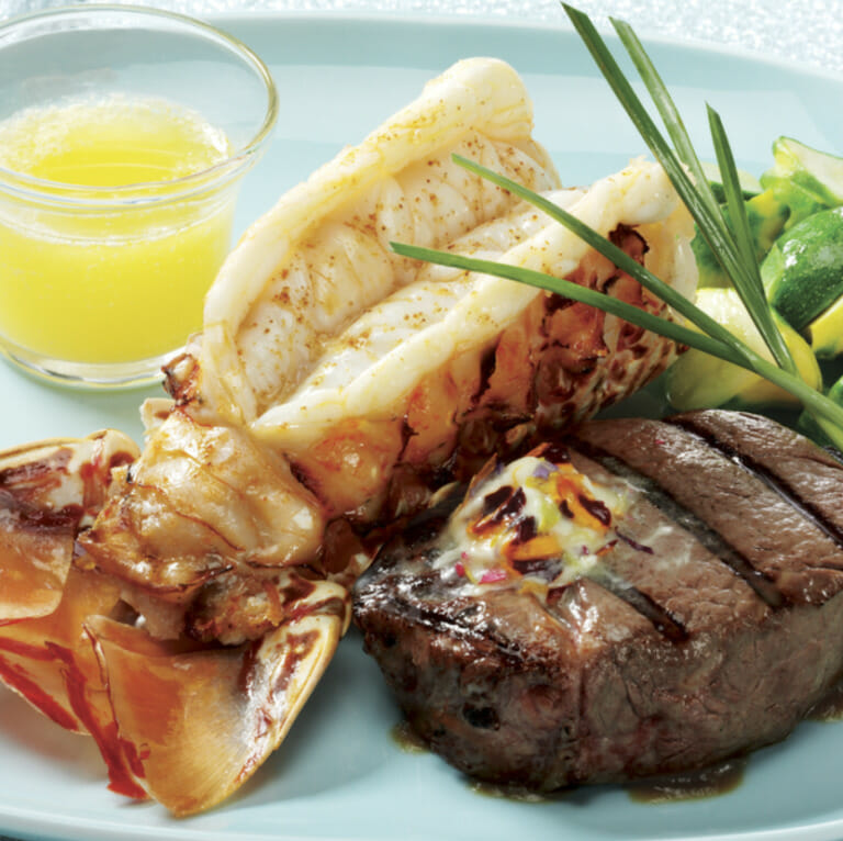 Grilled steak and lobster on a light blue plate with melted butter for dipping, brussel sprouts and green onions.