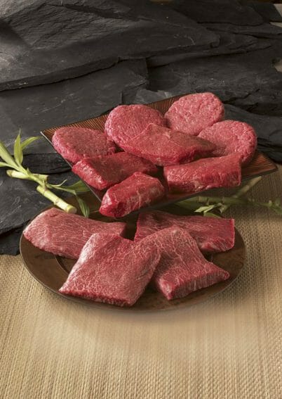 Two black plates of Kobe style beef steaks and burgers, ready to be cooked.