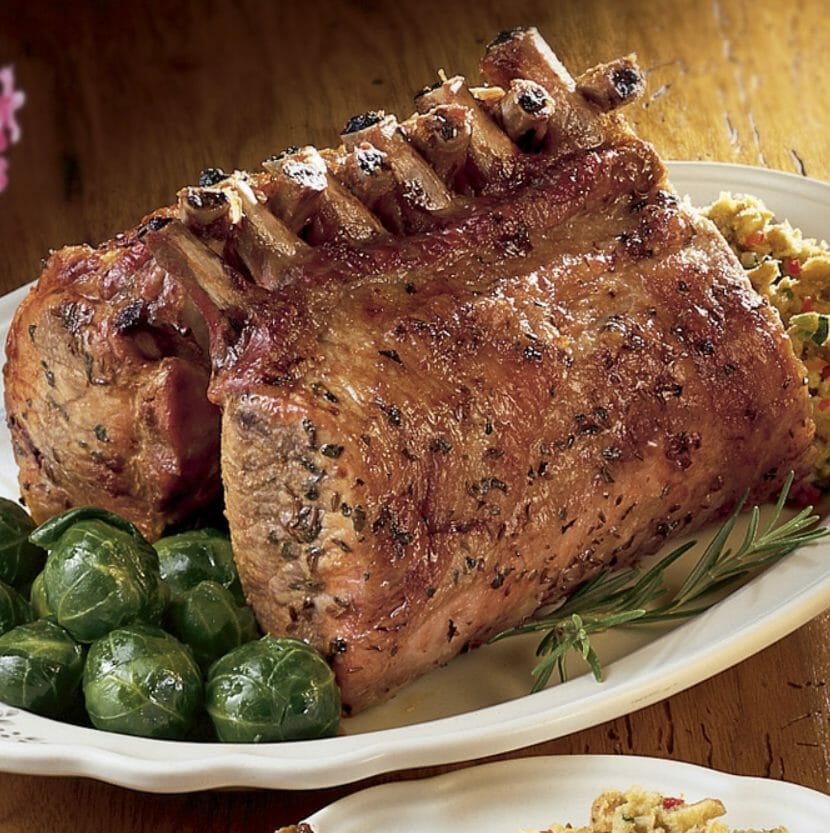 A rack of tasty seasoned pork resting on a white platter with buttered brussel sprouts, dressing, and a rosemary sprig.