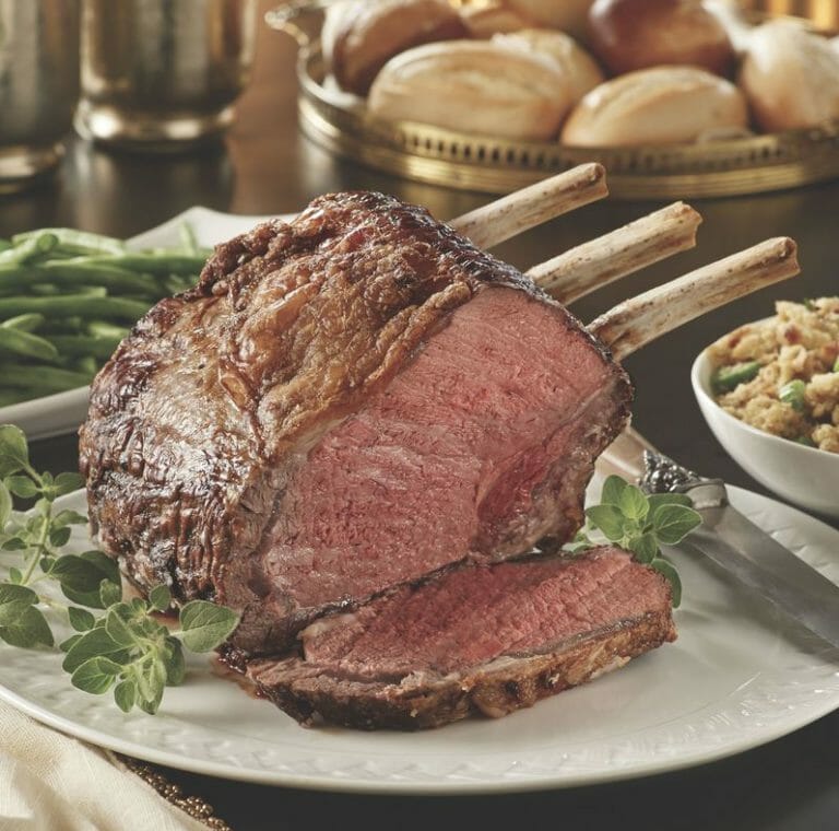 Medium-rare bone-in Prime Rib on a white platter, with bowls of green beans, dressing, and rolls on the table.
