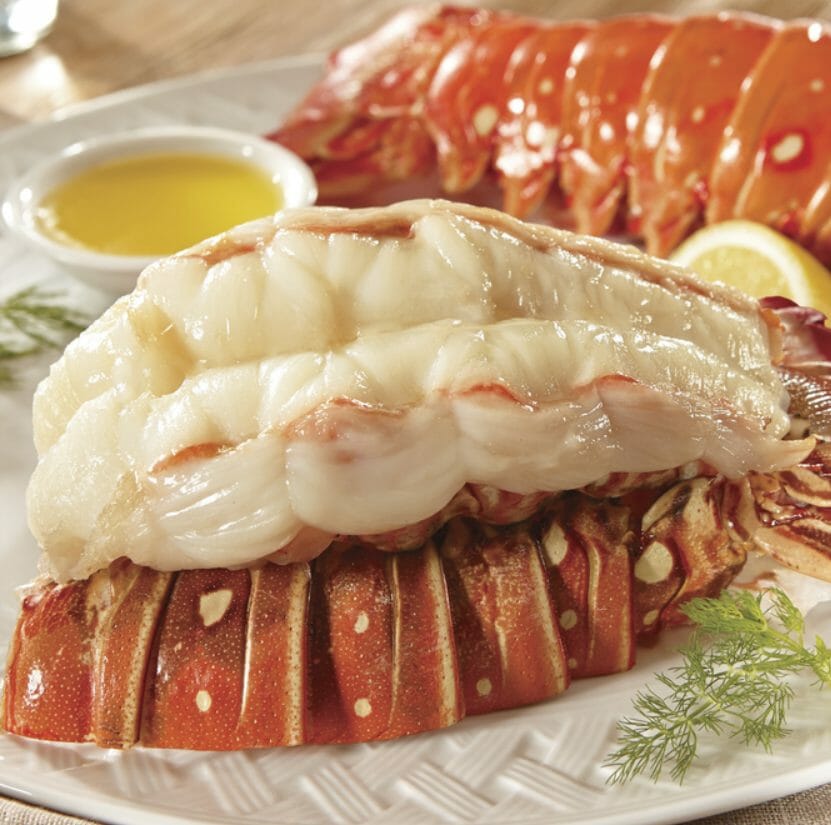 Two cooked lobster tails on a white plate with melted butter for dipping, a lemon wedge, and dill garnish.