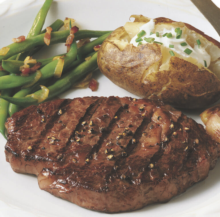 Seasoned, grilled Ribeye steak on a white plate with green beans and a baked potato with sour cream and chives.