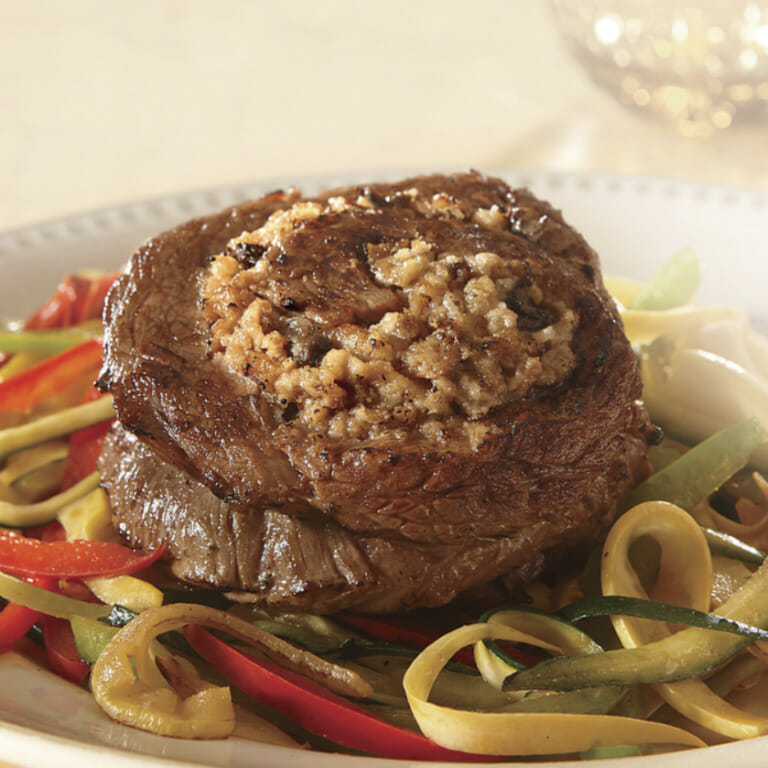 Two stuffed pin-wheel steaks on a bed of sauteed strips of onions, red peppers and squash on a white plate.
