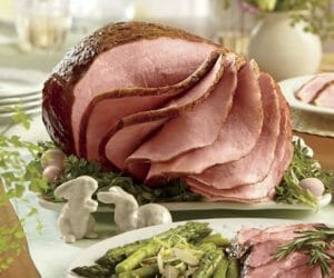 Our Masterpiece baked ham, sliced, on a bed of greens, with a nearby plate of ham, asparagus and rosemary garnish.