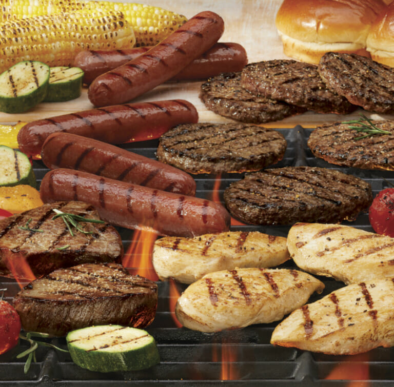 Grilled meats including burgers, chicken breasts, steaks, and hot dogs with grilled vegetables over a flaming grill.
