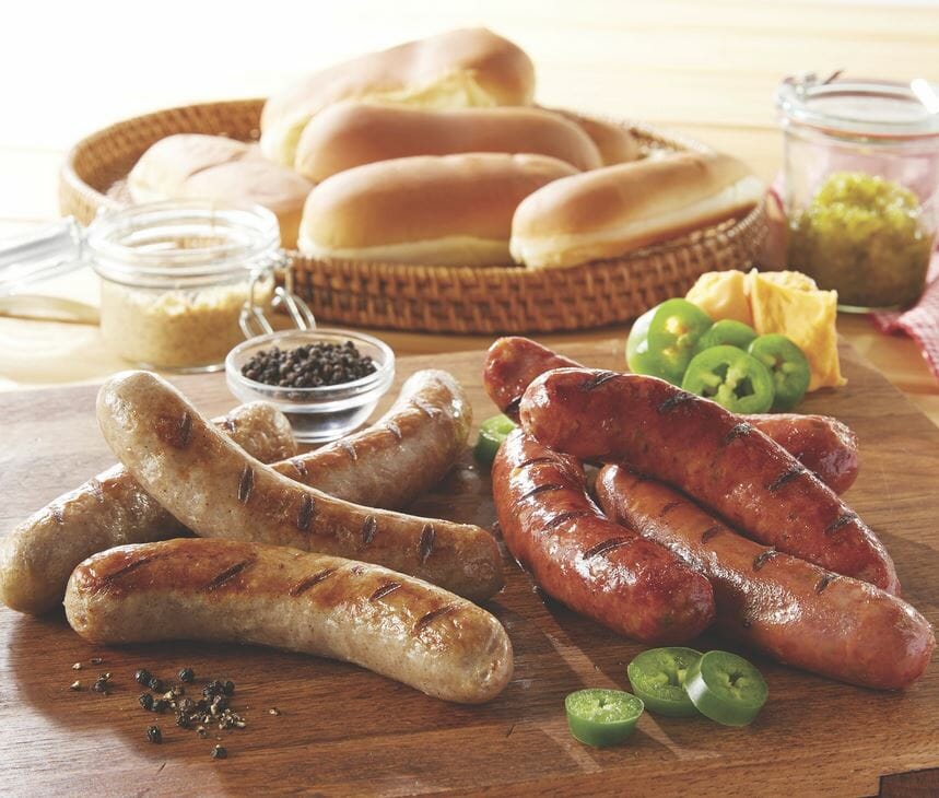 Two varieties of Bavarian Brats on a wooden platter, with sliced peppers, relish, mustard, and a basket of buns.