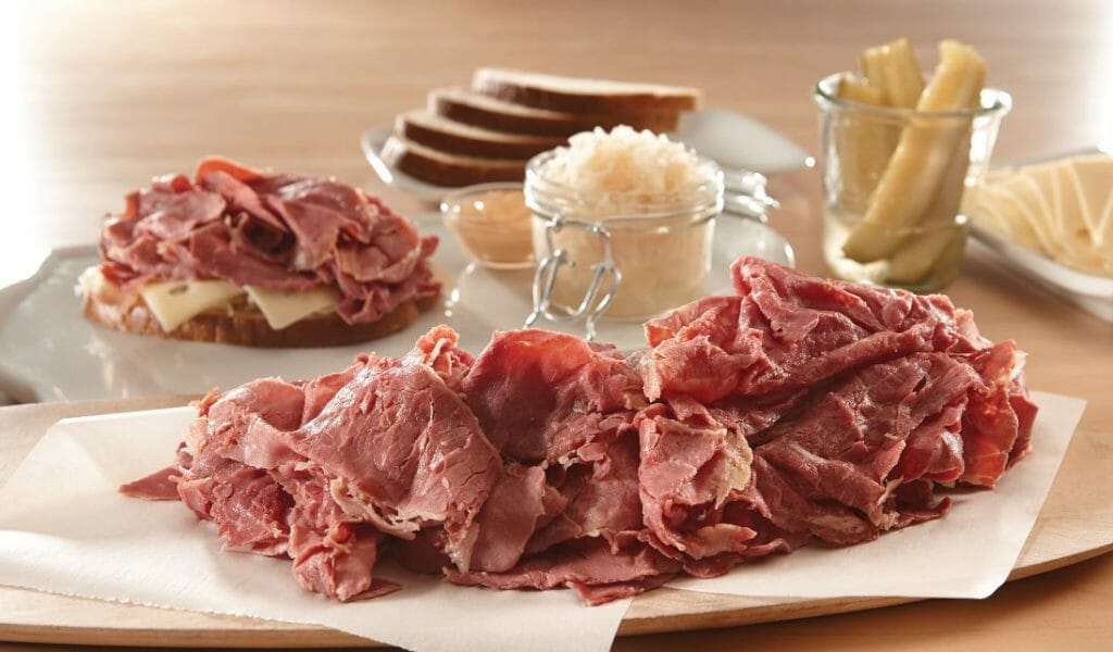 A pile of thinly sliced corned beef, with rye bread, saurkraut, Swiss cheese, dressing, and dill pickles for sandwiches.