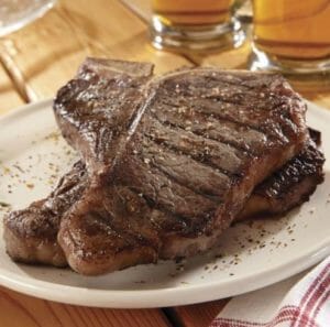 Two sizzling grilled and seasoned T-bone steaks on a white plate near glasses of ale on a wooden table.
