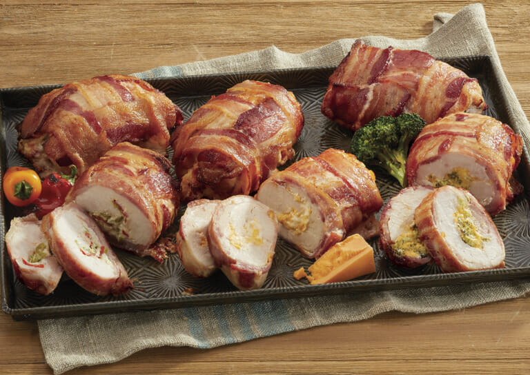 A platter of bacon wrapped stuffed chicken with broccoli, grilled peppers, and a cheese wedge for garnish.