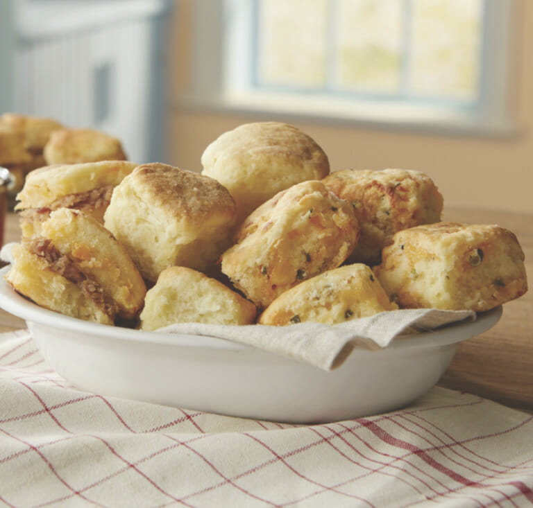A white bowl of herbed cheesy biscuits placed on a kitchen counter over a red and white linen towel.