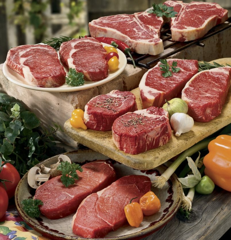 Raw VIP assortment of ten steaks on plates and platters with tomatoes, onions, mushrooms, parsley and garlic garnishes.