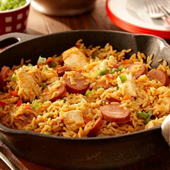 A cast iron pan hosting a Jambalaya mixture of seasoned rice, chicken, sausage and mixed vegetables on a dinner table.