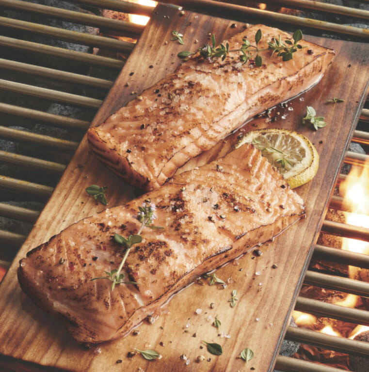 Two grilled salmon filets with a lemon wedge and thyme garnish on a wooden board placed over a grill with fiery coals.