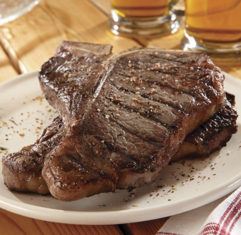 Two sizzling grilled and seasoned T-bone steaks on a white plate near glasses of ale on a wooden table.