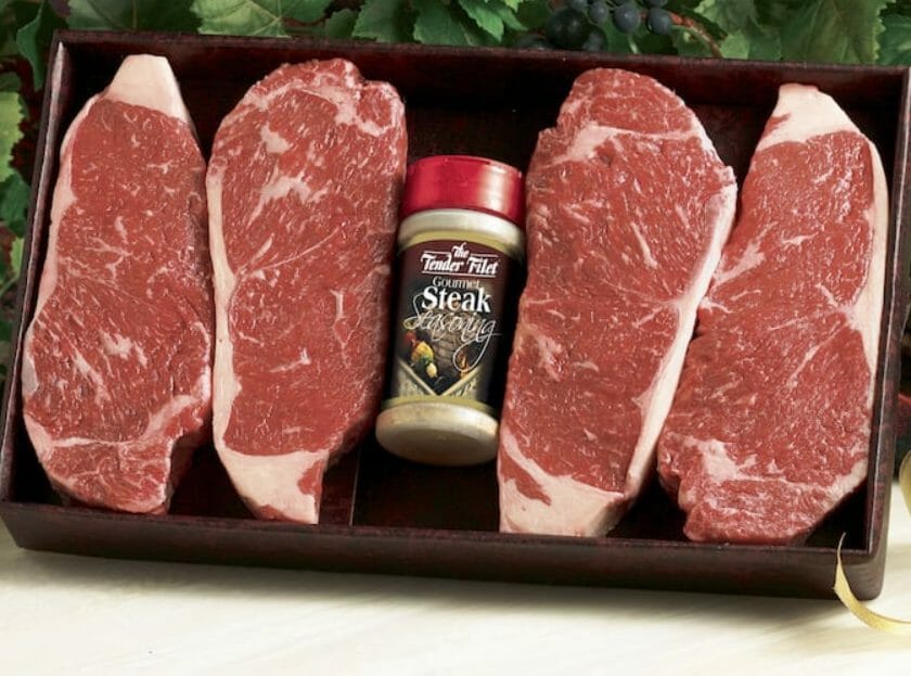 Four center cut New York Strip steaks with a bottle of Gourmet Seasoning in a pan, ready for grilling.