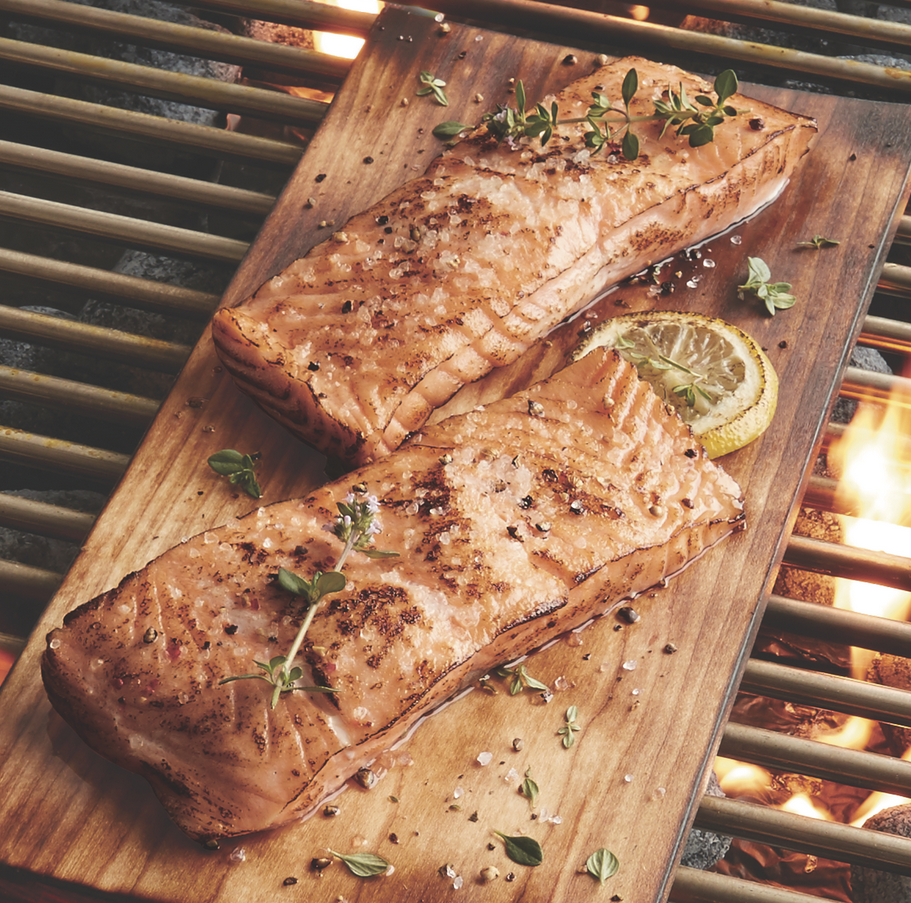 Two grilled salmon filets with a lemon wedge and thyme garnish on a wooden board placed over a grill with fiery coals.