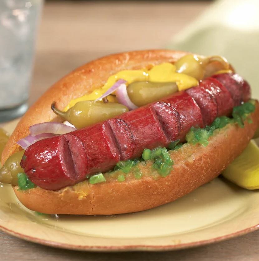 Angus beef frank on a bun with relish, peppers, onion and mustard, and dill pickle slices on the side of the plate.