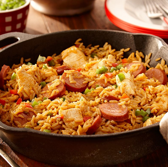 A cast iron pan hosting a Jambalaya mixture of seasoned rice, chicken, sausage and mixed vegetables on a dinner table.