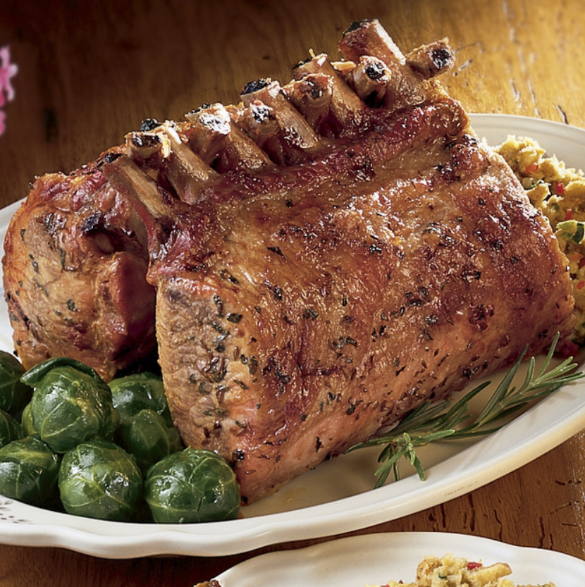 A rack of tasty seasoned pork resting on a white platter with buttered brussel sprouts, dressing, and a rosemary sprig.